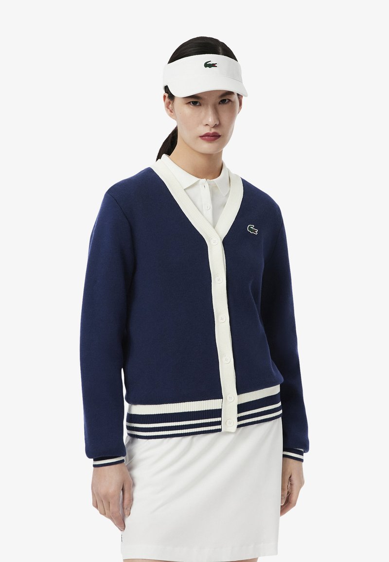 Cardigan in maglina blu navy con bottoni bianchi e polsini a coste; presenta un collo a V e un colletto a contrasto. Accentuato da un piccolo logo verde.