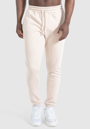 Pantalons de survêtement beige clair avec une taille élastique, cordon de serrage, coupe fuselée et poignets côtelés. Accompagnés de baskets blanches.