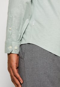 Jasnozielona koszula z długim rękawem, z mankietem na guziki, gładką fakturą oraz krojem, gdzie tył jest dłuższy. Pod spodem widoczna szara tkanina.