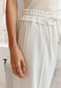 Witte broek met elastische tailleband en geplooide taille, voorzien van zijzakken en een zachte textuur. Een hand met een gouden ring is zichtbaar.