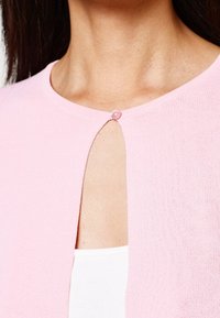 Chaqueta de punto rosa con cuello redondo, que presenta una pequeña abertura en la parte delantera asegurada con un botón rosa. Textura suave y tejido ligero.