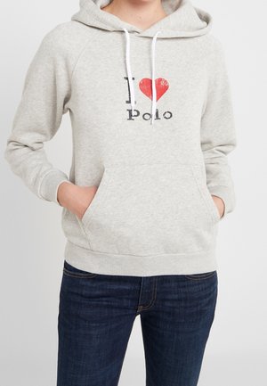 Osoba ubrana w jasnoszary kaptur z białymi sznurkami i czerwonym sercem z napisem "Kocham Polo", ręce w przedniej kieszeni, w połączeniu z ciemnoniebieskimi dżinsami.