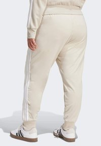 Béžové tepláky z mäkkého materiálu s tromi bielymi bočnými pruhmi, elastickým pásom a manžetami na členkoch, v kombinácii s bielymi teniskami.