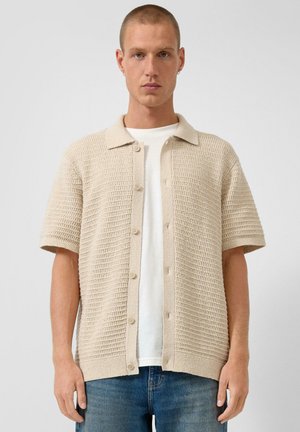 Homme portant une chemise beige à manches courtes en tricot avec des boutons sur un T-shirt blanc et un jean bleu, debout devant un fond uni.