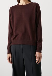 Femme portant un pull à manches longues bordeaux foncé et un pantalon ample noir, debout devant un fond clair uni.