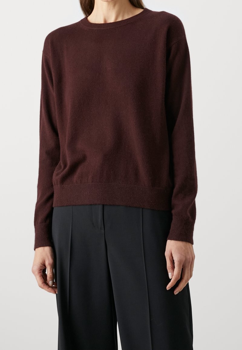 Femme portant un pull à manches longues bordeaux foncé et un pantalon ample noir, debout devant un fond clair uni.