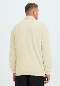 Beige Fleecejacke mit hohem Kragen, langen Ärmeln und einer weichen, strukturierten Oberfläche. Rückansicht zeigt einen geraden Saum und eine lockere Passform.