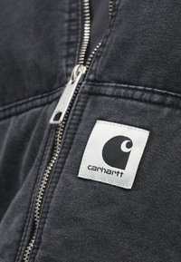 Melnā rāvējslēdzēja jaka ar teksturētu audumu, kuru rotā balta kvadrātveida etiķete ar melnu logotipu. Rāvējslēdzēja detaļa redzama gar šuvi.