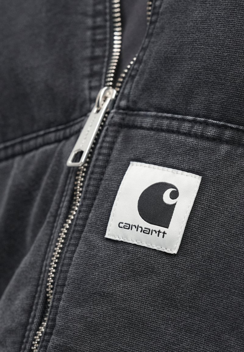 Zwarte jas met rits van gestructureerde stof, voorzien van een wit vierkant etiket met een zwart logo. Ritsdetail zichtbaar langs de naad.