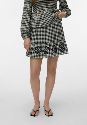 Femme portant une jupe à carreaux noire et blanche avec une broderie florale à l'ourlet, assortie à des sandales noires à brides sur un fond uni.