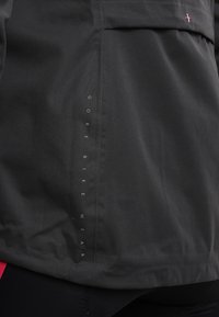 Chaqueta de Black Gore Bike Wear fabricada con tela que presenta texto de la marca en blanco de forma vertical y un pequeño detalle en cruz rosa cerca del bolsillo.