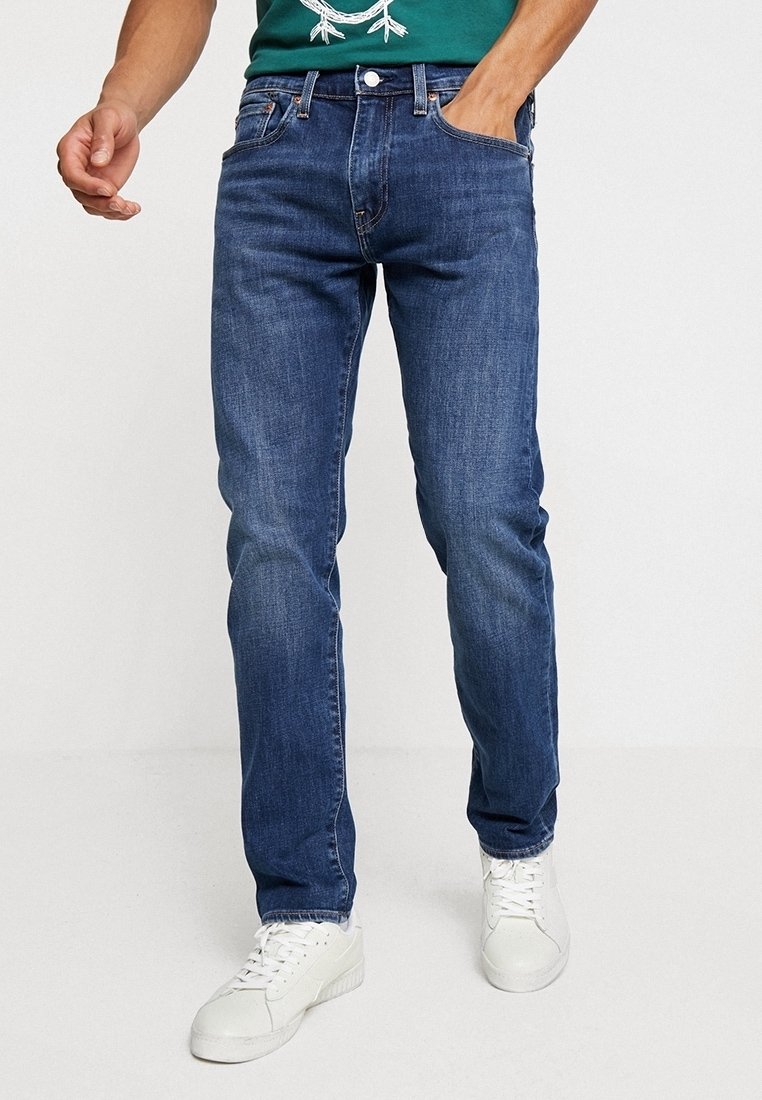 Homme portant un jean bleu slim, une chemise verte et des baskets blanches, une main dans la poche et en position de marche sur fond blanc.