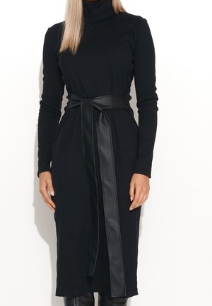 Femme portant une robe midi noire à manches longues et col roulé, avec une large ceinture en cuir nouée à la taille.