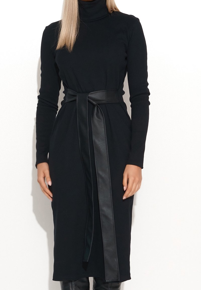 Femme portant une robe midi noire à manches longues et col roulé, avec une large ceinture en cuir nouée à la taille.