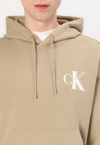 Smėlio spalvos džemperis su gobtuvu, pagamintas iš lygaus audinio, su priekiu kišene. Krūtinėje yra baltas "ck" logotipas ir reguliuojama raištelių sistema.