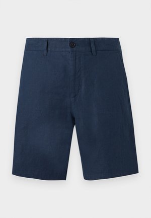 Homme portant une chemise beige à manches courtes boutonnée, un short bleu marine et des tongs noires, debout les mains dans les poches sur fond blanc.