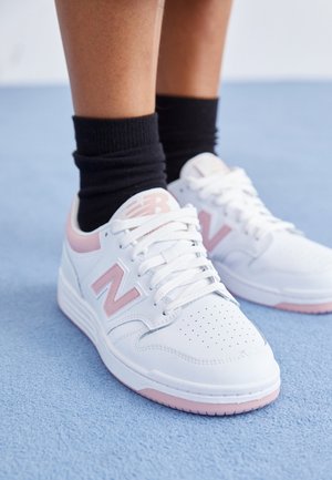 Personne portant des baskets New Balance blanches et roses avec des chaussettes noires, debout sur une surface bleu clair.