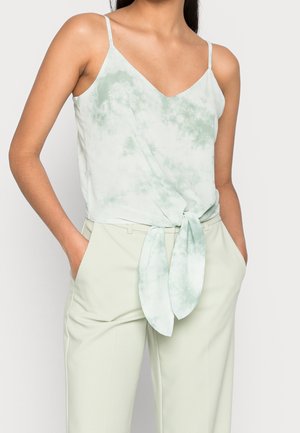 Femme portant un haut sans manches tie-dye vert clair avec un détail noué devant et un pantalon beige clair taille haute, mains dans les poches.