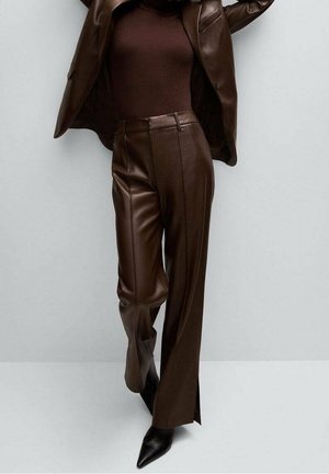 Person trägt einen braunen Lederblazer, ein braunes Oberteil, braune Lederhose und schwarze spitze Schuhe und steht vor einem schlichten hellen Hintergrund.