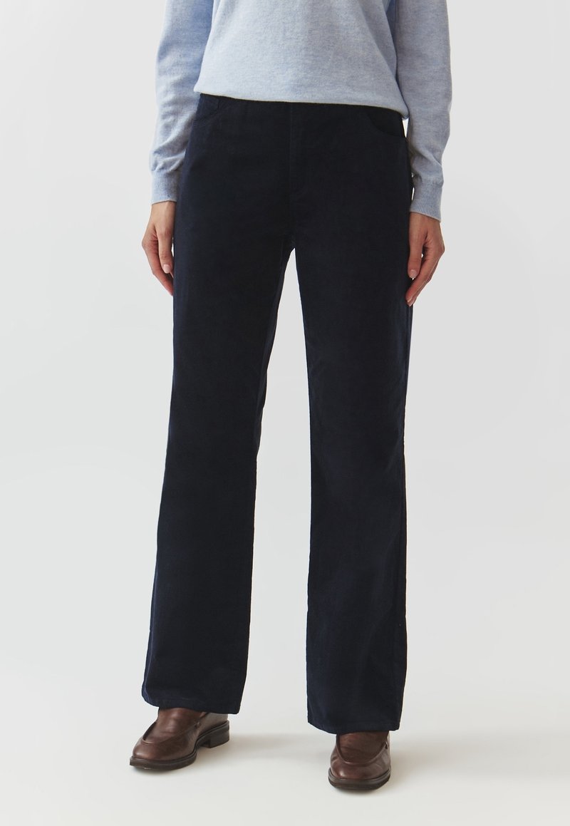 Personne portant un pantalon large bleu marine foncé, un haut à manches longues gris clair et des chaussures en cuir marron, debout devant un fond uni.