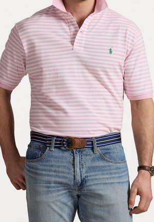 Mann trägt pink-weiß gestreiftes Poloshirt mit grünem Logo, blaue Jeans, gestreiften Gürtel mit Lederschnalle und Armbanduhr am rechten Arm.