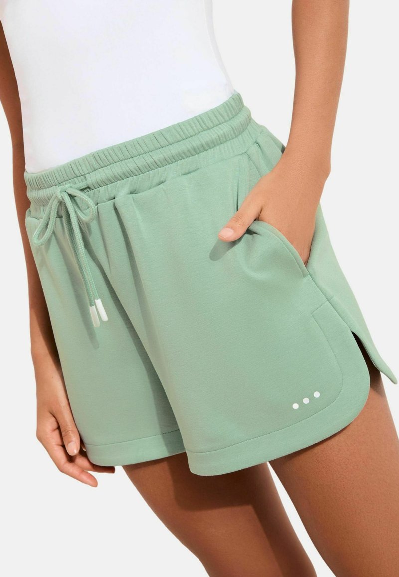 Pantaloni scurți atletici de culoare verde deschis, realizați dintr-un material moale, prevăzuți cu talie elastică cu șnur, buzunare laterale și detalii decorative de cusături.