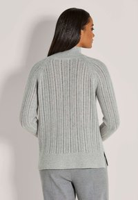 Frau mit dunklem, glattem Haar, die einen hellgrauen, gerippten Strickpullover und graue Hose trägt, mit dem Rücken zu einer schlichten Hintergrund.