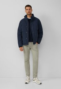 Navy Blouson mit Stehkragen, ausgestattet mit Druckknöpfen, kombiniert mit hellgrünen Hose und beigen Sneakern mit grauen Akzenten.