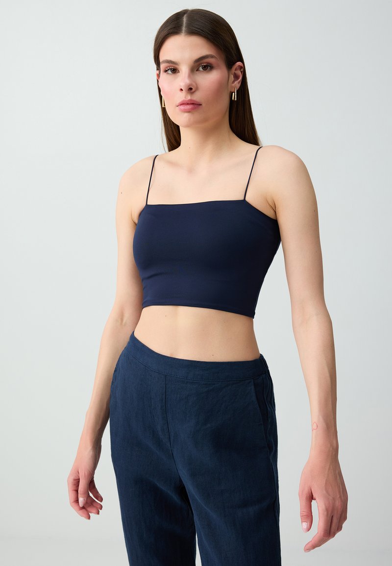 Jeune femme aux longs cheveux bruns portant un crop top sans manches bleu marine et un pantalon taille haute sur un fond uni.