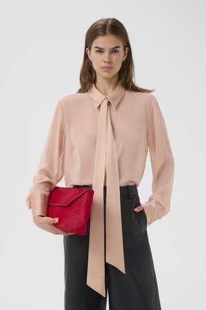 Žena v svetloružovej blúzke s dlhým kravatovým uzlom, čiernych nohaviciach, držiaca červenú listovú kabelku v ľavej ruke na jednoduchom pozadí.