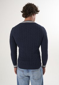 Maglione blu navy a trecce con un motivo testurizzato, orlo e polsini a costine, e colletto a strisce bianche. Indossato con jeans chiari.