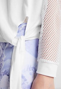 Weißes Sweatshirt mit Knoten-Detail an der Taille, mit Netzärmeln und glatter Stofftextur. Darunter hellblaue, gemusterte Leggings.