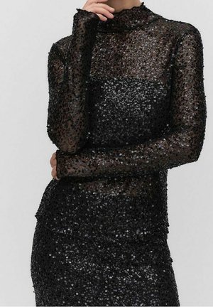 Kobieta w dopasowanej, długorękawowej czarnej sukience ozdobionej czarnymi cekinami, z przezroczystym materiałem na ramionach i górnej części klatki piersiowej.