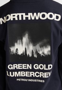 Camicia a maniche lunghe di colore blu navy con una grande grafica in bianco e nero di alberi su uno sfondo cupo, con testo bianco audace sopra e sotto.