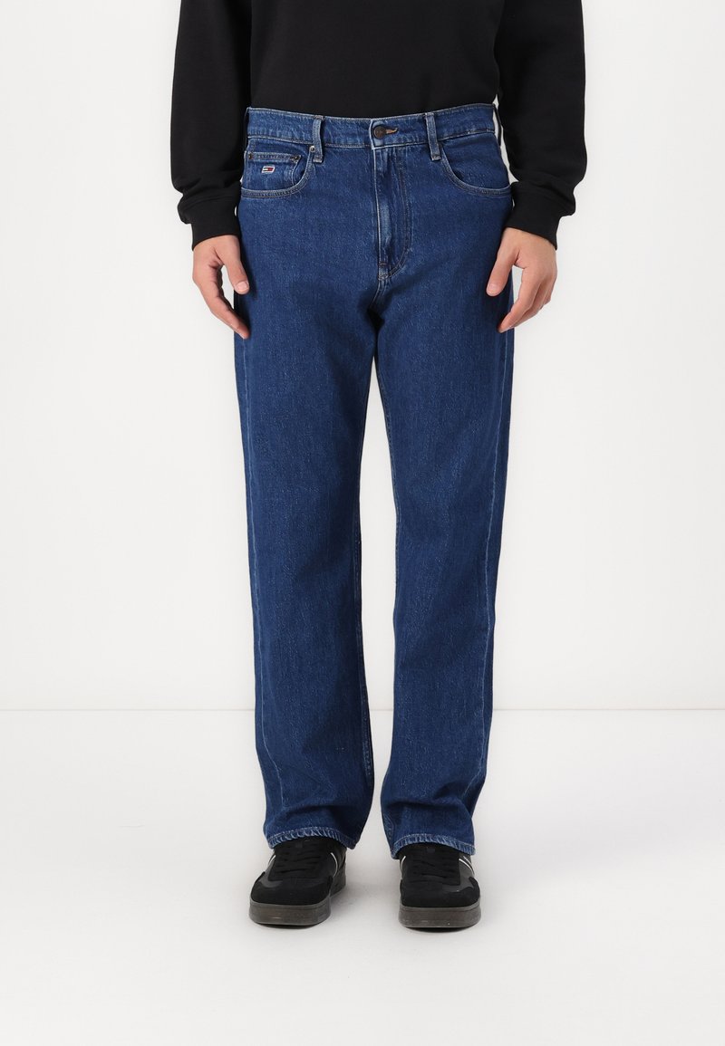 Blue denim jeans with a straight fit, mid-rise waist, five-pocket design, and contrast stitching. Worn with black sneakers.