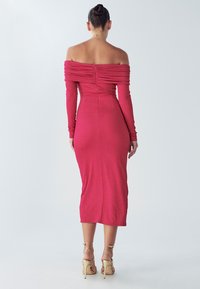 Midi šaty s odhalenými ramenami v jasne ružovej látke, s dlhými rukávmi a nariasenou textúrou. Zapínanie na zips na chrbte. Béžové sandále na remienkoch.