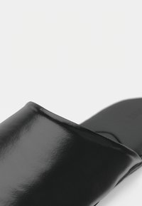 Close-up de uma sandália deslize preta brilhante com uma faixa larga sobre o pé e uma sola plana em um fundo branco.