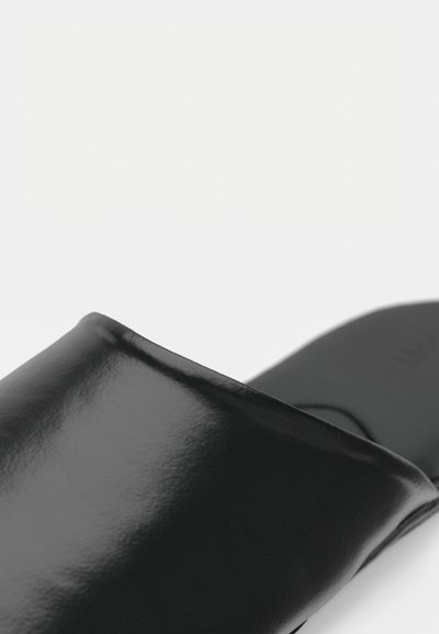 Gros plan sur une sandale noire brillante avec une large sangle sur le pied et une semelle plate sur fond blanc.