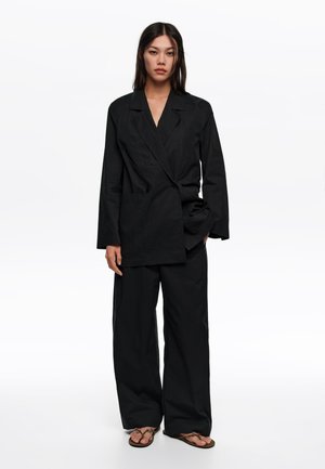 Mujer con un traje negro suelto de pantalones de pierna ancha y una chaqueta cruzada, combinados con sandalias marrones, de pie sobre un fondo blanco.