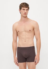 Young man with light skin and short brown hair wearing dark brown boxer briefs, standing against a plain white background.