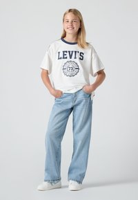 Djevojka stoji s rukama u džepovima, nosi bijelu Levi's majicu, svijetloplave traperice i bijele tenisice, ispred jednostavne pozadine.