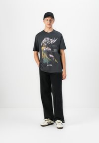 Young man wearing black cap, black Marvel graphic tee, wide-leg black pants, and white Adidas sneakers, standing against a plain white background.