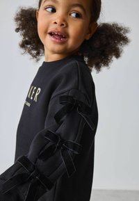 Felpa nera con scritte dorate, caratterizzata da maniche lunghe decorate con fiocchi in velluto sulla cucitura esterna, realizzata in morbido cotone e con scollo rotondo.