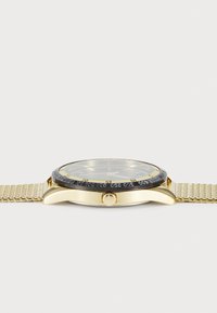 Montre dorée avec un bracelet en maille, lunette noire avec des chiffres en relief, et un cadran rond affichant des accents verts et un verre clair.
