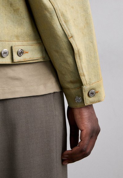 Light green denim jacket with silver snap buttons, orange stitching, and cuff details, worn over a beige shirt and gray trousers.