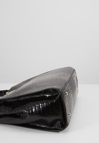 Sac à main en cuir synthétique effet crocodile noir avec des clous dorés aux coins inférieurs, posé sur le côté sur une surface blanche.