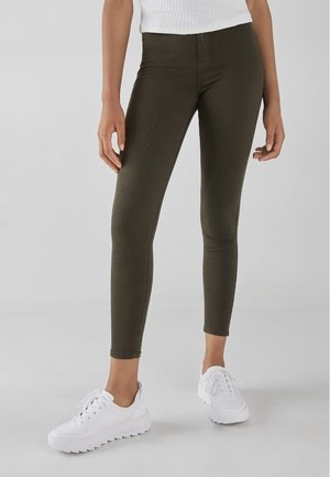 Personne portant un pantalon skinny vert olive foncé, un haut blanc côtelé et des baskets blanches épaisses à lacets, debout devant un fond clair uni.