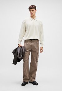 Ανοιχτό κρέμα μακρυμάνικο πουκάμισο πόλο, μπεζ παντελόνι cargo με τσέπες και καφέ δερμάτινο σακάκι. Μαύρα παπούτσια ολοκληρώνουν το σύνολο.