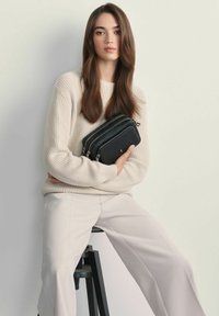 Młoda kobieta z długimi brązowymi włosami, ubrana w beżowy sweter dzianinowy i spodnie, siedząca na stołku, trzymająca czarną torebkę na zamek.