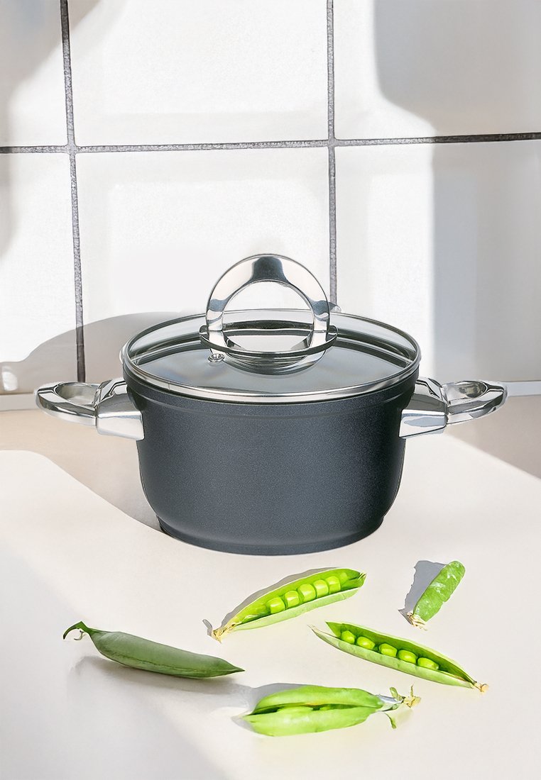 Černý hrnec na vaření se skleněnou poklicí a kovovými držadly na kuchyňské lince, obklopený otevřenými a zavřenými lusky zeleného hrášku.
